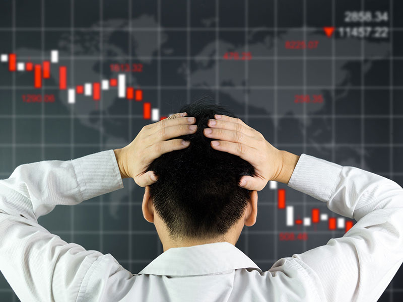 Homme d'affaire de dos, les mains sur la tête, devant un tableau montrant des indices boursiers à la baisse.