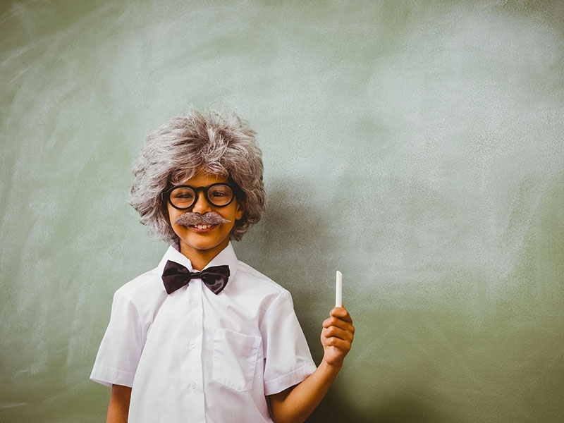 Enfant déguisé en petit Einstein, se tenant debout devant un tableau d'école, une craie à la main.
