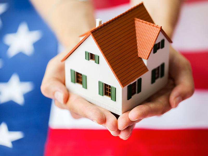 Petite maison tenue dans les mains d'une jeune femme, avec drapeau américain en arrière-plan.