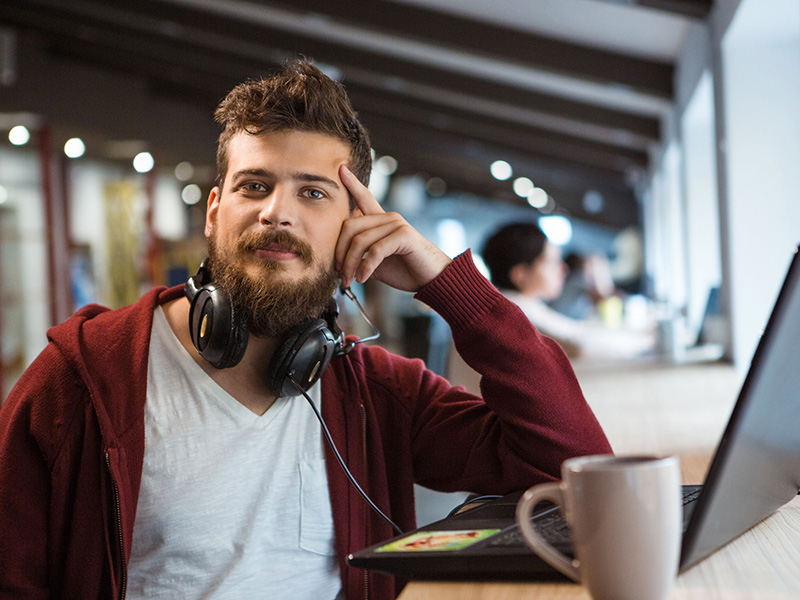 Jeune homme confiance, avec barbe et écouteurs, assis devant son ordinateur portable.