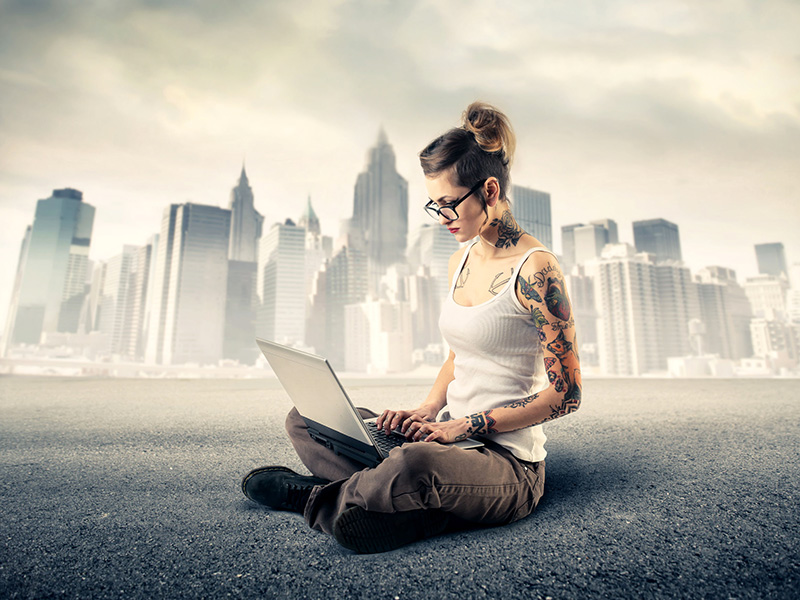 Jeune femme avec tatouages, assise par terre et travaillant sur un ordinateur portable.