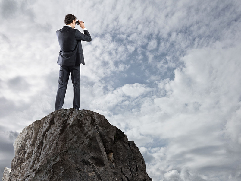 Homme d'affaires debout sur un pic rocheux, scrutant l'horizon avec des jumelles.