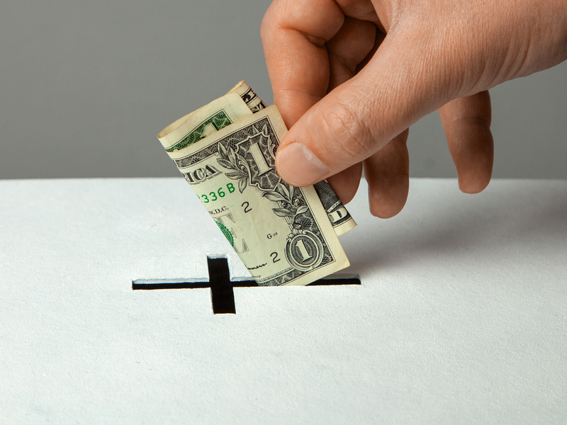 Main qui dépose un dollar dans une boîte dont l'ouverture est en forme de croix.