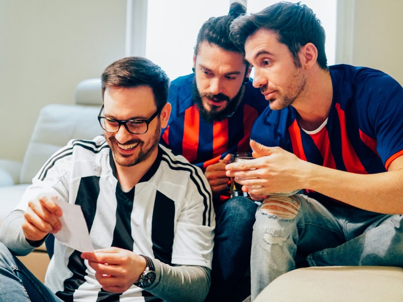 Trois hommes en maillot sportif dans un salon regardent un document de pari sportif.