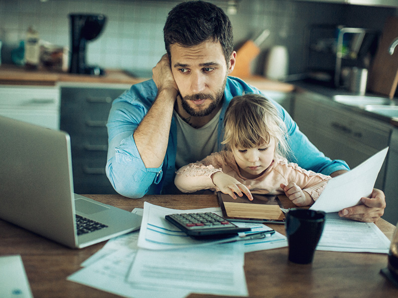 Un père semblant soucieux, tenant dans ses bras un jeune enfant, devant un ordinateur portable et une calculatrice.