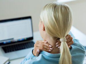 Une femme assise devant un ordinateur sur un bureau. Elle se masse le cou.
