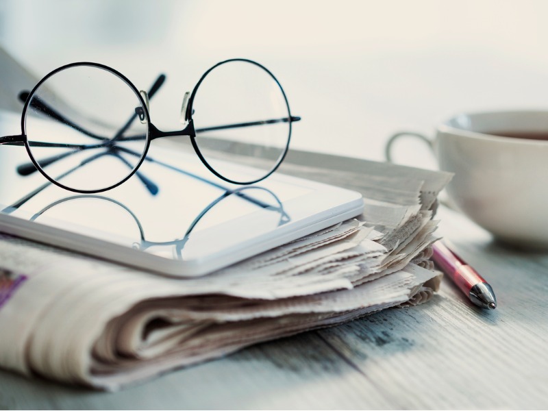 Une paire de lunettes posée sur un calepin, posé sur un journal, posé sur un bureau à côté d'un stylo et d'un café.