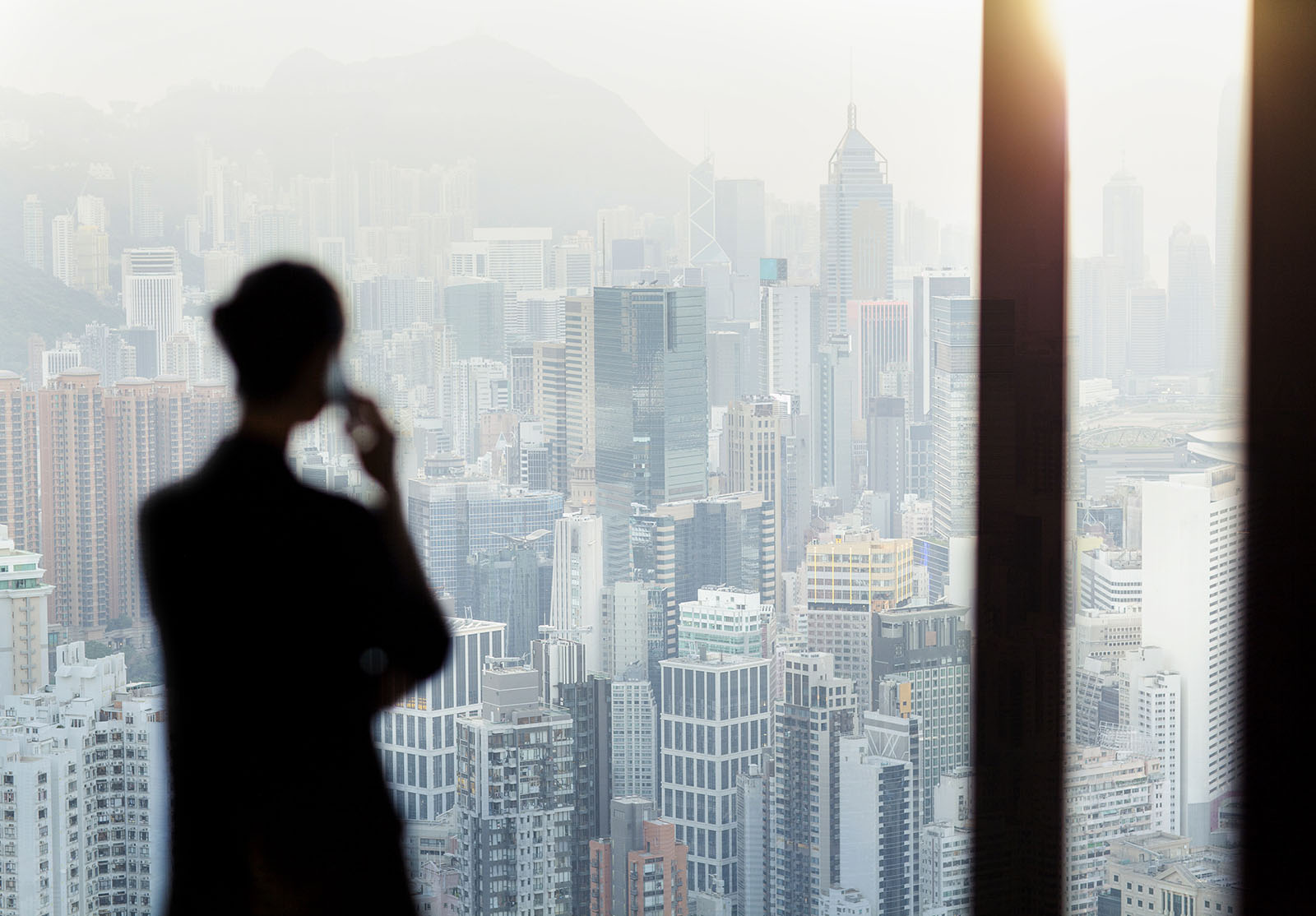 Femme d'affaires parlant au téléphone depuis la fenêtre de son bureau, avec vue sur le paysage urbain de Hong Kong.