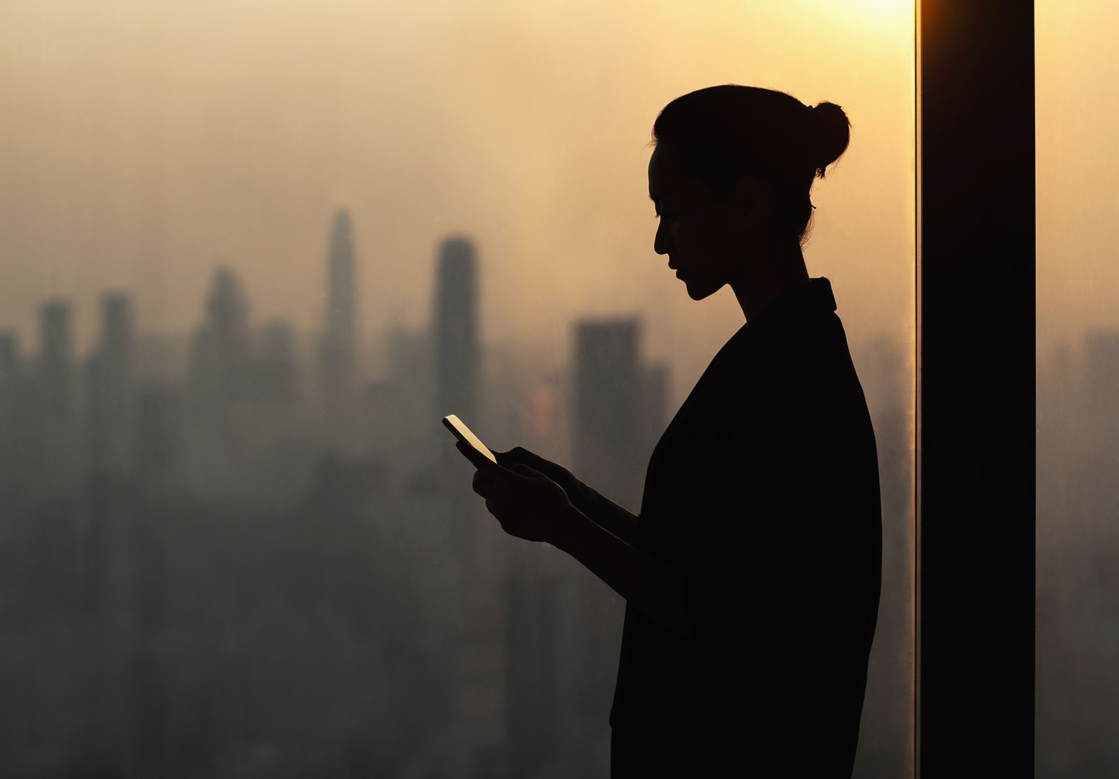 Silhouette d'une jeune femme utilisant un smartphone à côté d'une fenêtre avec un paysage urbain, Shenzhen, Chine