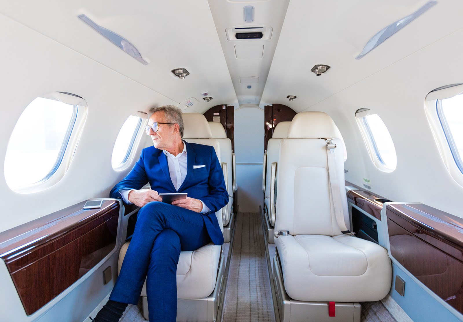 Homme d'affaires en costume, assis dans un avion privé, tenant une tablette numérique, regardant par le hublot.