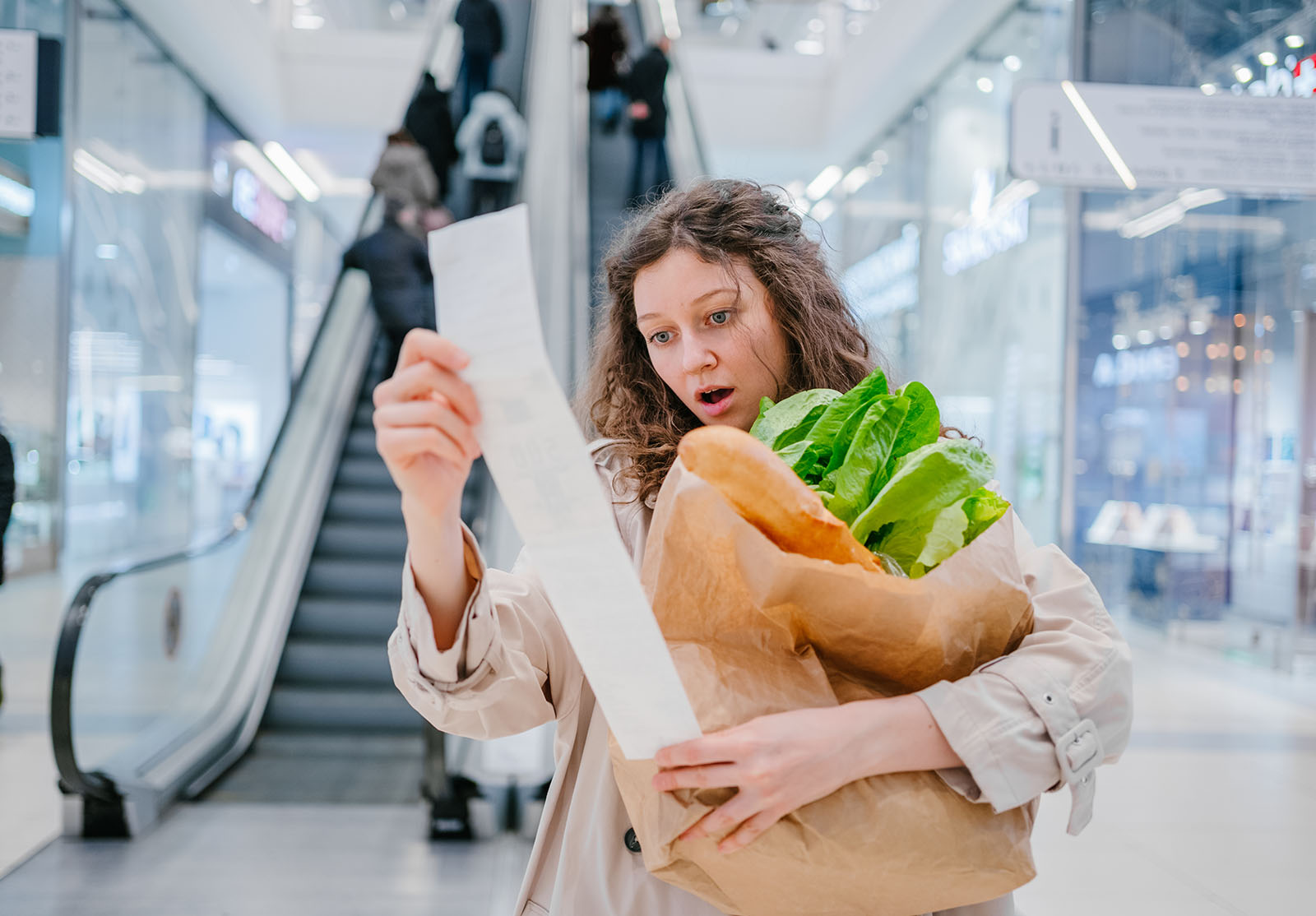 Une femme en état de choc regarde un chèque en papier d'un supermarché dans un centre commercial sur fond d'escalator et tient un paquet avec des produits frais, une augmentation des prix.