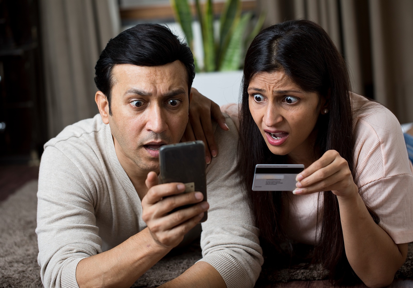 Couple choqué qui a l’air inquiet en utilisant un téléphone portable et une carte de crédit à la maison.