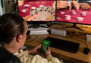Un homme assis avec son téléphone dans la main devant deux écrans montrant du poker en ligne.