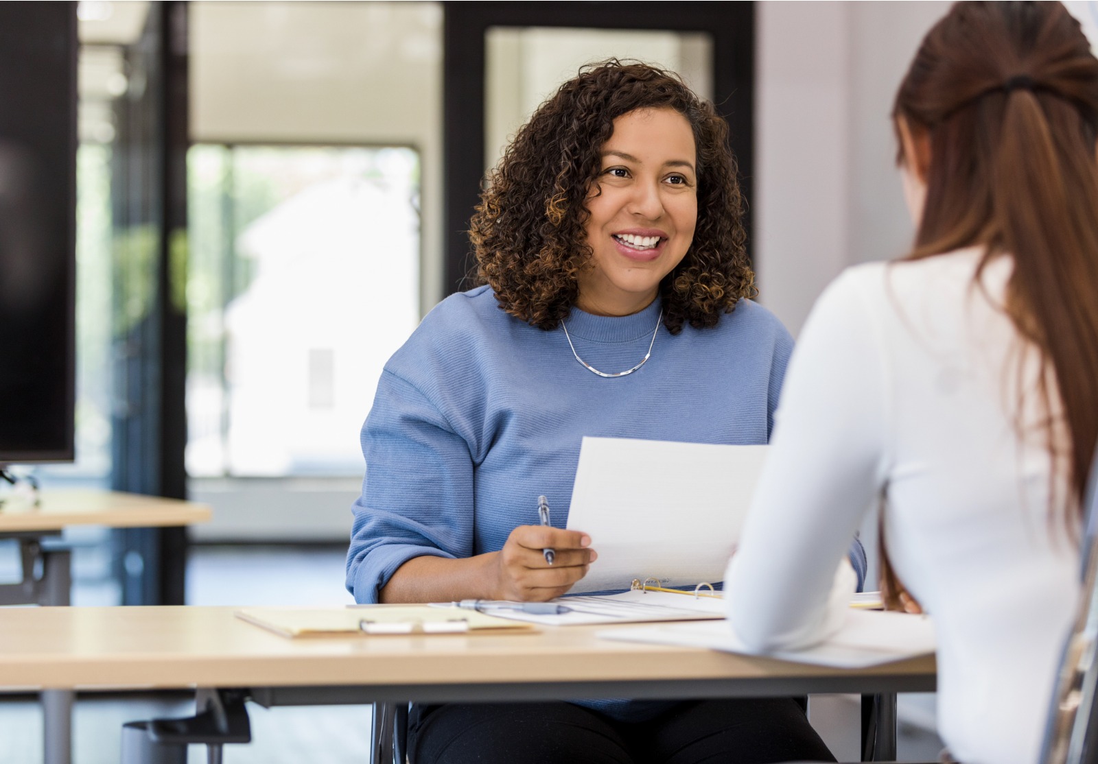 Une femme souriante tenant des feuilles et un stylo derrière un bureau parle à une autre femme.